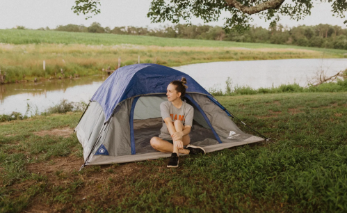 A person sits outside a tent by a calm river, surrounded by green grass and trees.
