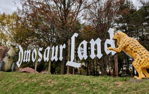 Sign for "Dinosaur Land" with a yellow leopard statue beside it, surrounded by trees and autumn foliage.