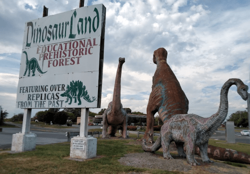 Sign for Dinosaur Land featuring large dinosaur replicas in a prehistoric forest setting under a cloudy sky.