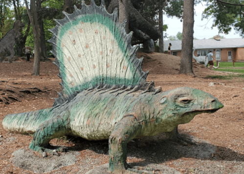 A large, colorful dinosaur sculpture with a spiked back, set in a wooded area with people in the background.