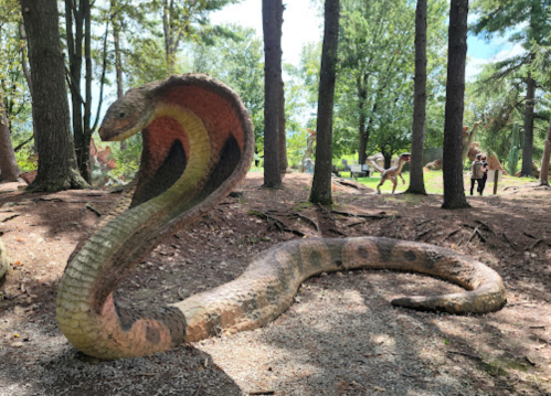 A large, colorful snake sculpture in a wooded area, with people and dinosaur sculptures in the background.