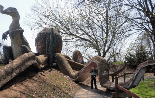 A person stands next to a large, colorful dinosaur sculpture and a winding slide in a park setting.