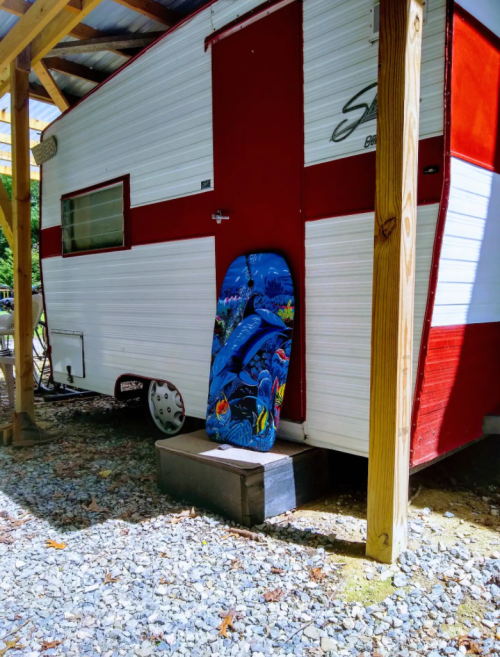 A red and white camper trailer with a colorful surfboard propped against it, surrounded by gravel and wooden posts.