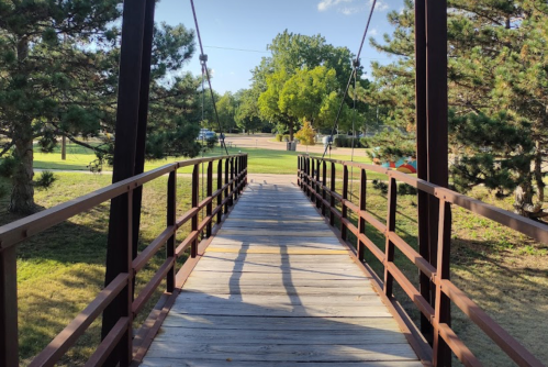 A wooden bridge with a metal frame spans a grassy area, surrounded by trees and a clear blue sky.