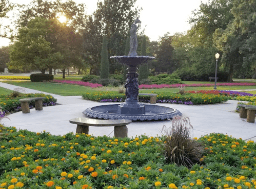 A serene garden scene featuring a fountain surrounded by colorful flowers and benches in a park setting.