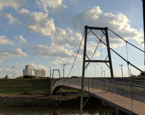 A suspension bridge spans a river, with a grassy bank and buildings in the background under a partly cloudy sky.