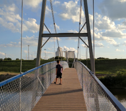 A person walks on a wooden bridge with a chain-link railing, surrounded by green grass and a cloudy sky.