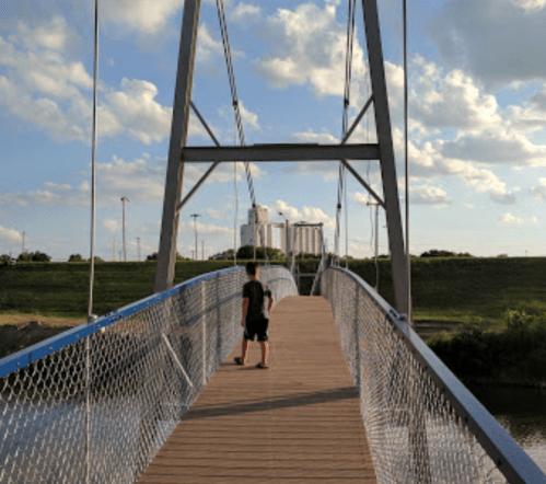 A person walks on a wooden bridge with a chain-link railing, surrounded by green grass and a cloudy sky.
