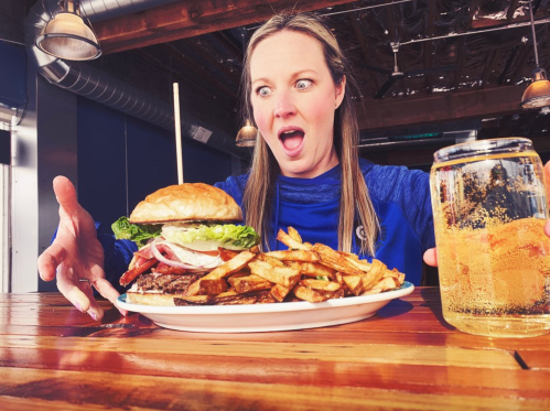 A surprised woman stares at a large burger and fries on a plate, with a drink beside her on a wooden table.