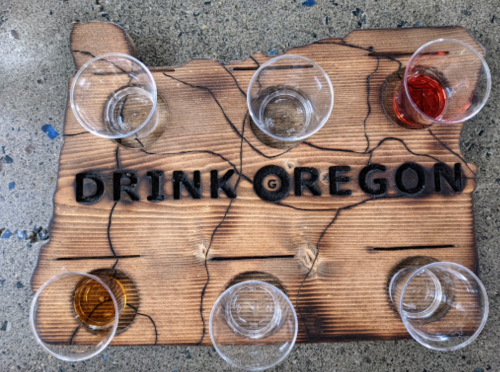 A wooden tasting board with the text "DRINK OREGON" and six small glasses filled with different beverages.