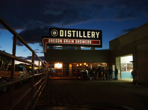 A distillery sign illuminated at night, with people gathered outside and a darkening sky in the background.