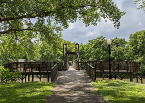 A wooden bridge leads to a gazebo surrounded by lush greenery and trees under a cloudy sky.