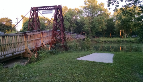 A rustic metal bridge over a calm waterway, surrounded by lush greenery and trees in a peaceful park setting.