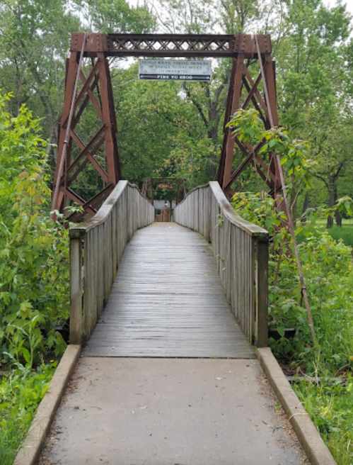 A wooden bridge with a metal frame surrounded by greenery, leading into a park area.