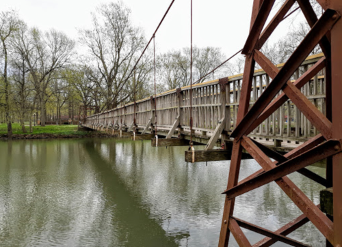 A wooden suspension bridge spans a calm river, surrounded by trees and greenery on a cloudy day.