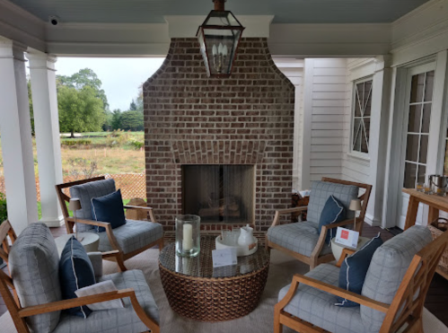 Cozy outdoor seating area with a brick fireplace, wooden chairs, and a round table, surrounded by greenery.