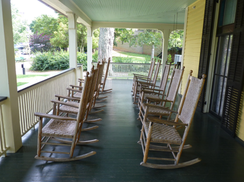A row of wooden rocking chairs on a porch, surrounded by greenery and a peaceful outdoor setting.