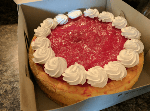 A round cheesecake topped with red fruit glaze and decorative white whipped cream rosettes, inside a cardboard box.