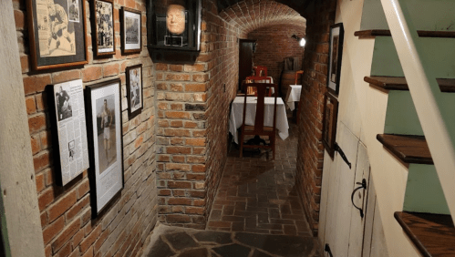 A dimly lit brick hallway with framed photos on the walls, leading to a dining area with wooden furniture.