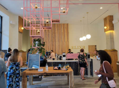 A modern café interior with a wooden counter, pink accents, and customers ordering drinks. Some people are wearing masks.