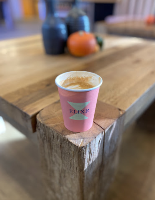 A pink coffee cup with the logo "ELIXR" sits on a wooden table, with decorative items in the background.