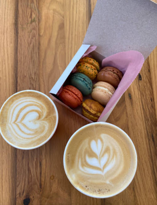 A box of colorful macarons next to two cups of latte art on a wooden table.