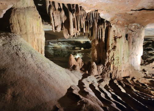A dimly lit cave interior featuring stalactites and stalagmites, with textured rock formations and shadows.