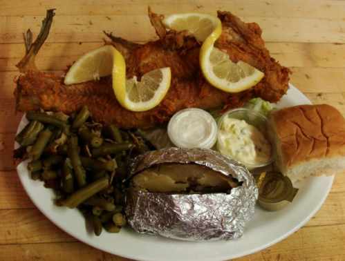 A plate featuring fried fish with lemon, green beans, a baked potato in foil, coleslaw, and a dinner roll.