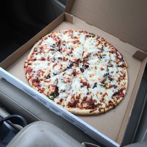 A large pizza with cheese and toppings in a pizza box, resting on the seat of a car.