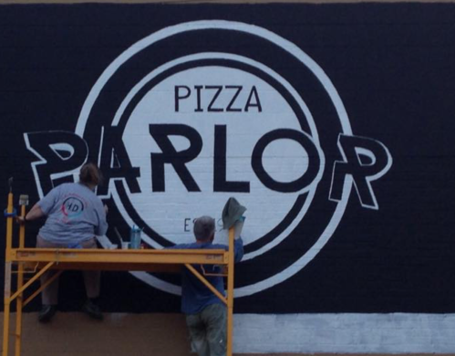 Two people painting a large mural that reads "Pizza Parlor" on a black wall.