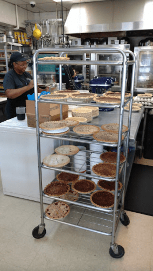 A metal cart filled with various pies, with a person preparing food in the background of a kitchen.