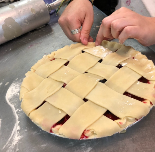 A person weaving a lattice crust over a pie filled with red fruit, on a stainless steel countertop.