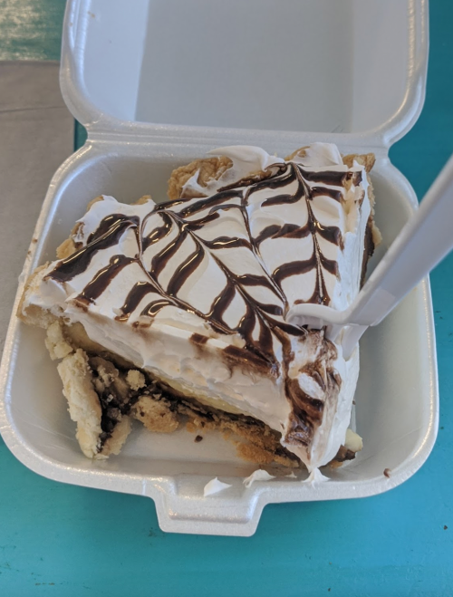 A slice of pie with a creamy topping and chocolate drizzle, served in a takeout container with a plastic fork.