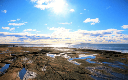 A rocky shoreline under a bright blue sky with scattered clouds and sunlight reflecting on the water.