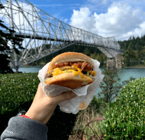 A hand holding a burger with bacon and cheese, with a bridge and river in the background under a partly cloudy sky.