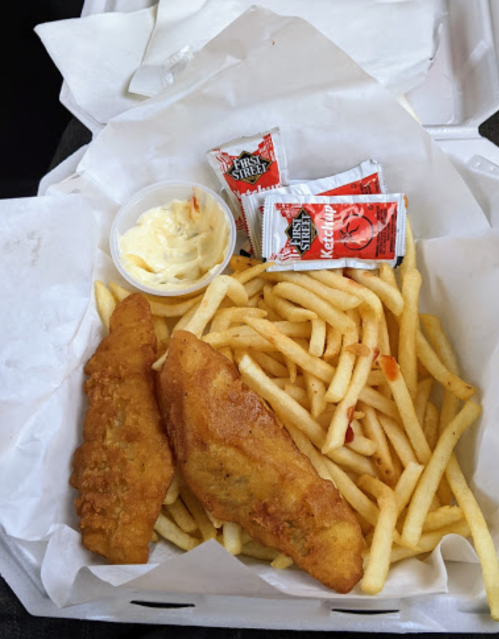 A takeout box with two pieces of fried fish, a portion of fries, and condiments including ketchup and mayonnaise.