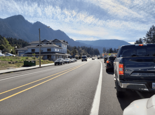 A busy road lined with cars, mountains in the background, and a building on the left under a blue sky.