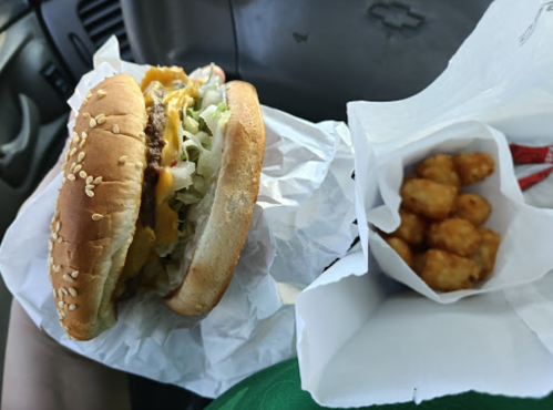 A sesame seed bun burger with lettuce and cheese next to a small container of tater tots in a car setting.