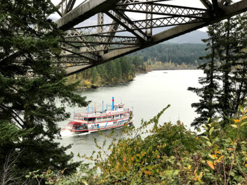 A riverboat navigates a calm river, framed by trees and a bridge overhead, with autumn foliage in the background.