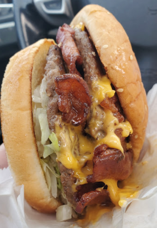 A close-up of a juicy burger with two beef patties, melted cheese, crispy bacon, and lettuce in a sesame seed bun.