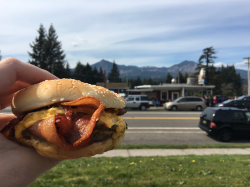 A hand holding a bacon cheeseburger with mountains and a diner in the background on a sunny day.
