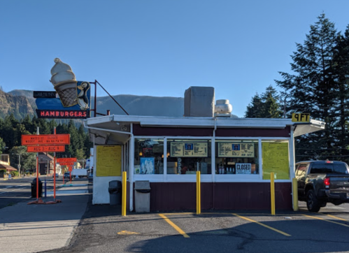 A small hamburger stand with a large ice cream cone sign, surrounded by trees and mountains in the background.