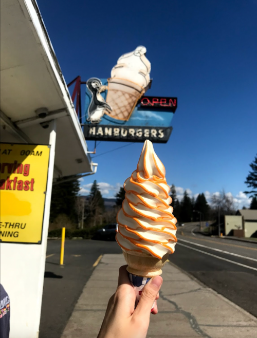 A hand holds a soft-serve ice cream cone with orange swirl, in front of a diner sign featuring an ice cream cone.