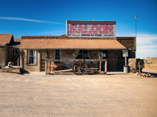 A rustic wooden saloon with a large sign, set in a desert landscape under a clear blue sky.