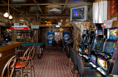 A rustic bar interior with a pool table, slot machines, and a TV, featuring wooden decor and beer advertisements.