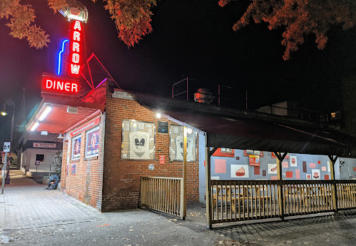 A brightly lit diner at night with a neon sign, featuring outdoor seating and colorful wall art.