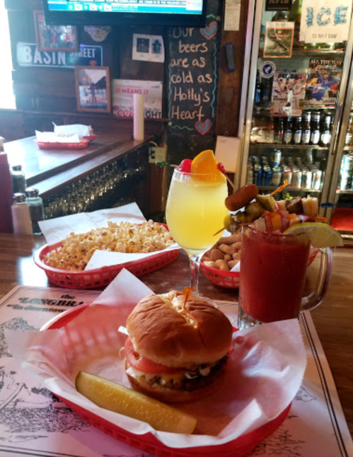 A bar scene featuring a burger with a pickle, a colorful cocktail, and snacks like popcorn and skewered appetizers.