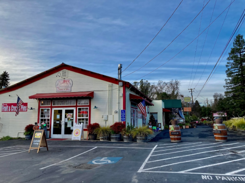 A quaint store with a red roof, American flags, and outdoor seating, surrounded by trees and a clear blue sky.