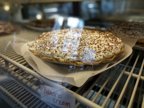 A banana cream pie with a whipped topping and chocolate shavings, displayed in a glass case.