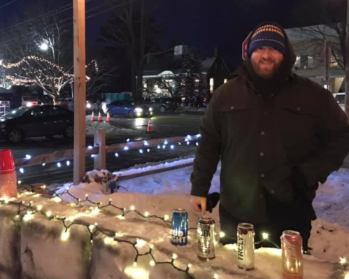 A man in a winter coat stands by a snowy ledge with colorful lights and several cans of beer in front of him.